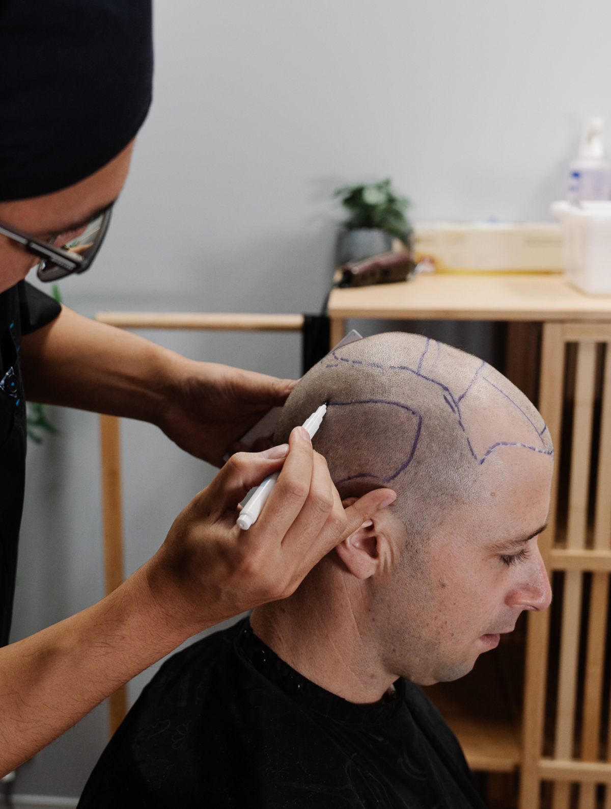 A person with a shaved head is seated while another person wearing glasses and a hat uses a marker to draw guidelines on the scalp. The background features wooden shelves with a few items on top. The atmosphere appears to be a salon or barbershop.
