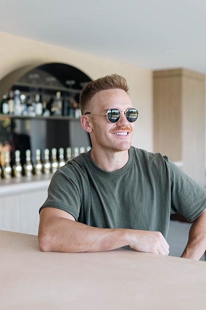 A man with short hair wearing sunglasses and a casual olive green t-shirt is smiling while sitting at a bar counter. Behind him is a well-stocked bar with various bottles and a mirror. The setting appears clean and modern.