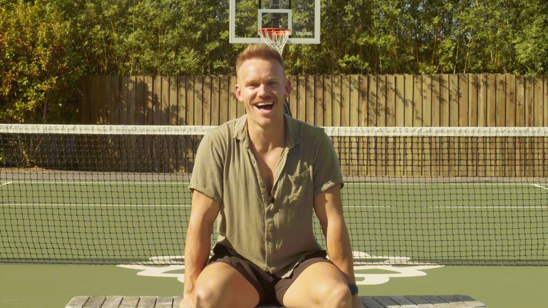 A man is sitting on a wooden bench on an outdoor basketball court. He is smiling and laughing, wearing a short-sleeved, button-up shirt and shorts. The net and hoop are visible behind him, with greenery and a wooden fence in the background.