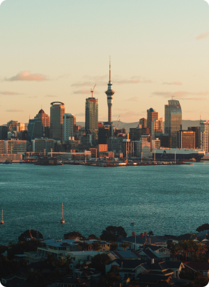 A scenic view of a city skyline with a prominent tower at the center, surrounded by various skyscrapers. The buildings are illuminated by the soft light of a sunrise or sunset. In the foreground, there is a body of water and rooftops of smaller buildings.
