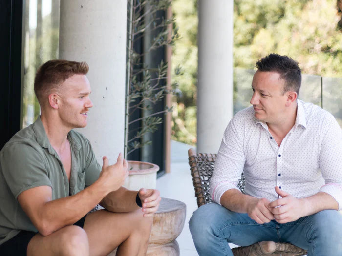 Two men are engaged in a conversation on a covered porch. One man, wearing a green short-sleeved shirt and shorts, gestures with his hand while speaking. The other man, in a white polka-dot shirt and jeans, listens attentively. Trees are visible in the background.