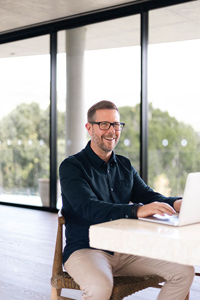 A man with glasses and a beard, wearing a dark shirt and light pants, is sitting on a wooden chair, smiling and typing on a laptop. He is in a bright room with large windows and a view of trees outside.